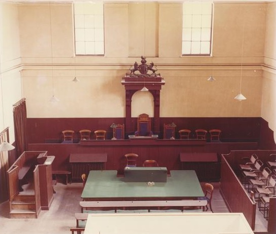 Faded colour photo of Court 2 - the Magistrates Court. Dark coloured wooden Magistrates bench with Royal Arms above.