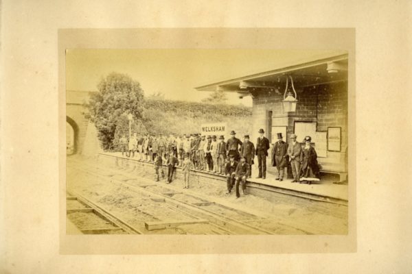 Sepia photograph of people dressed in suits standing on a railway platform.