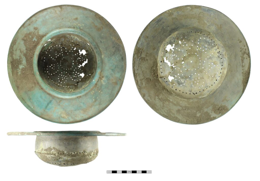 A Roman bronze strainer, seen from above, below and from the side. This is a patinated green deep bowl with a very broad flat rim. The base of the bowl is perforated with holes in a flower-like pattern.