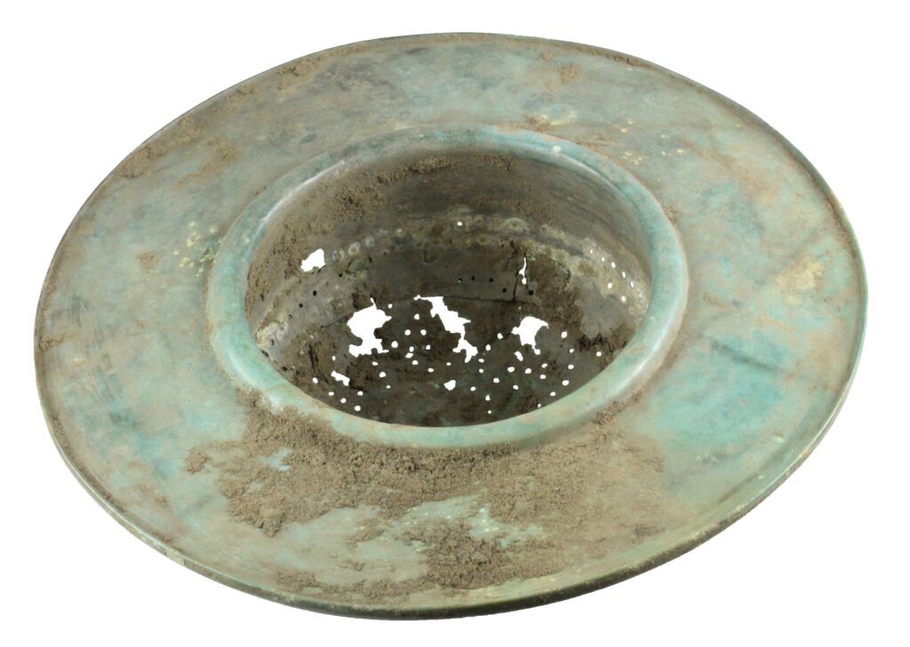 A Roman bronze strainer, seen from one side. This is a patinated green deep bowl with a very broad flat rim. The base of the bowl is perforated with holes in a flower-like pattern. There are clear traces of wear and damage in the base of the bowl.