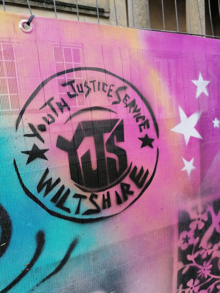 A graffiti piece of pink and blue. The Youth Justice Service Wiltshire logo is stamped in the centre.