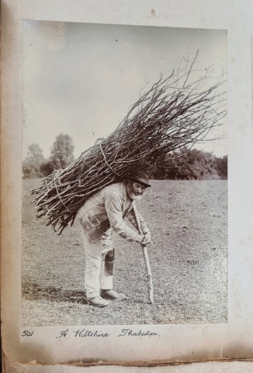 Black and white photograph of an old man, carrying a bundle of sticks on his back. A grey beard underlining his weathered face, the figure stoops whilst apparently pausing for the photographer, his leathery hands grasp the pole supporting the bundle of hazel on his back.